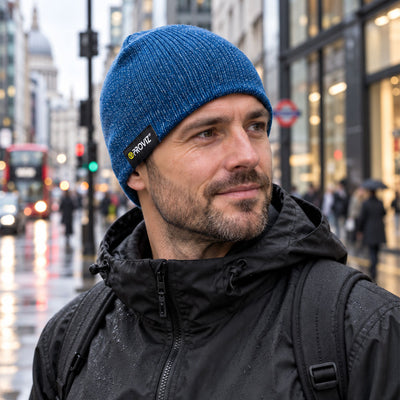 lifestyle A man wearing a blue heathered knit beanie with a black brand tab that reads 'Proviz' and a black hooded jacket, photographed outdoors on a city street with blurred buildings and pedestrians in the background.
