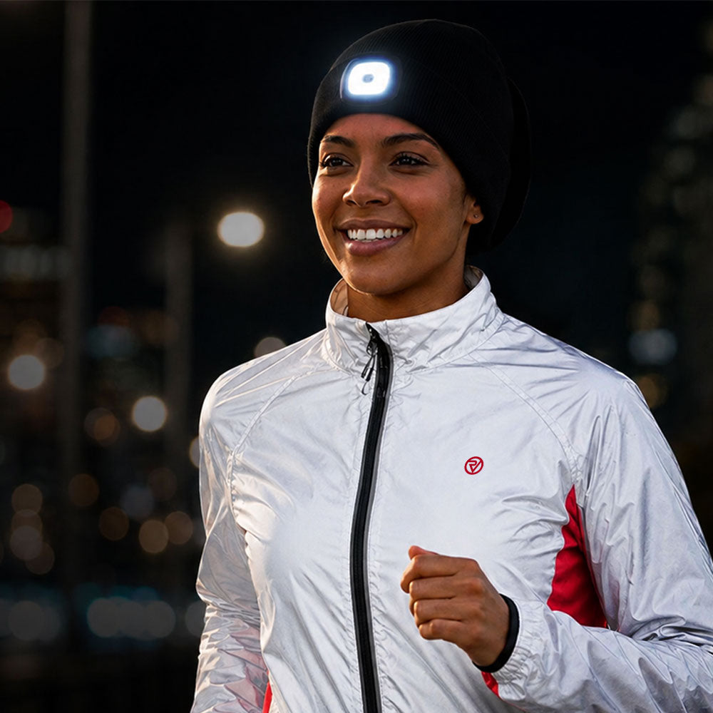 A smiling woman wearing a black beanie with a bright white LED light on the front, a white zip-up athletic jacket with red side panels, and a black wristband, photographed outdoors at night with blurred city lights in the background. Lifestyle photo