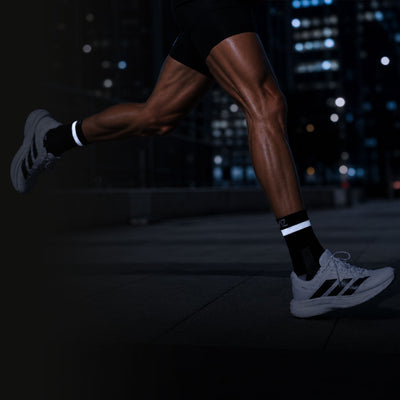 lifestyle A runner's legs in black athletic shorts, Proviz black socks with white stripes, and white running shoes with black stripes, captured at night against a blurred cityscape background.