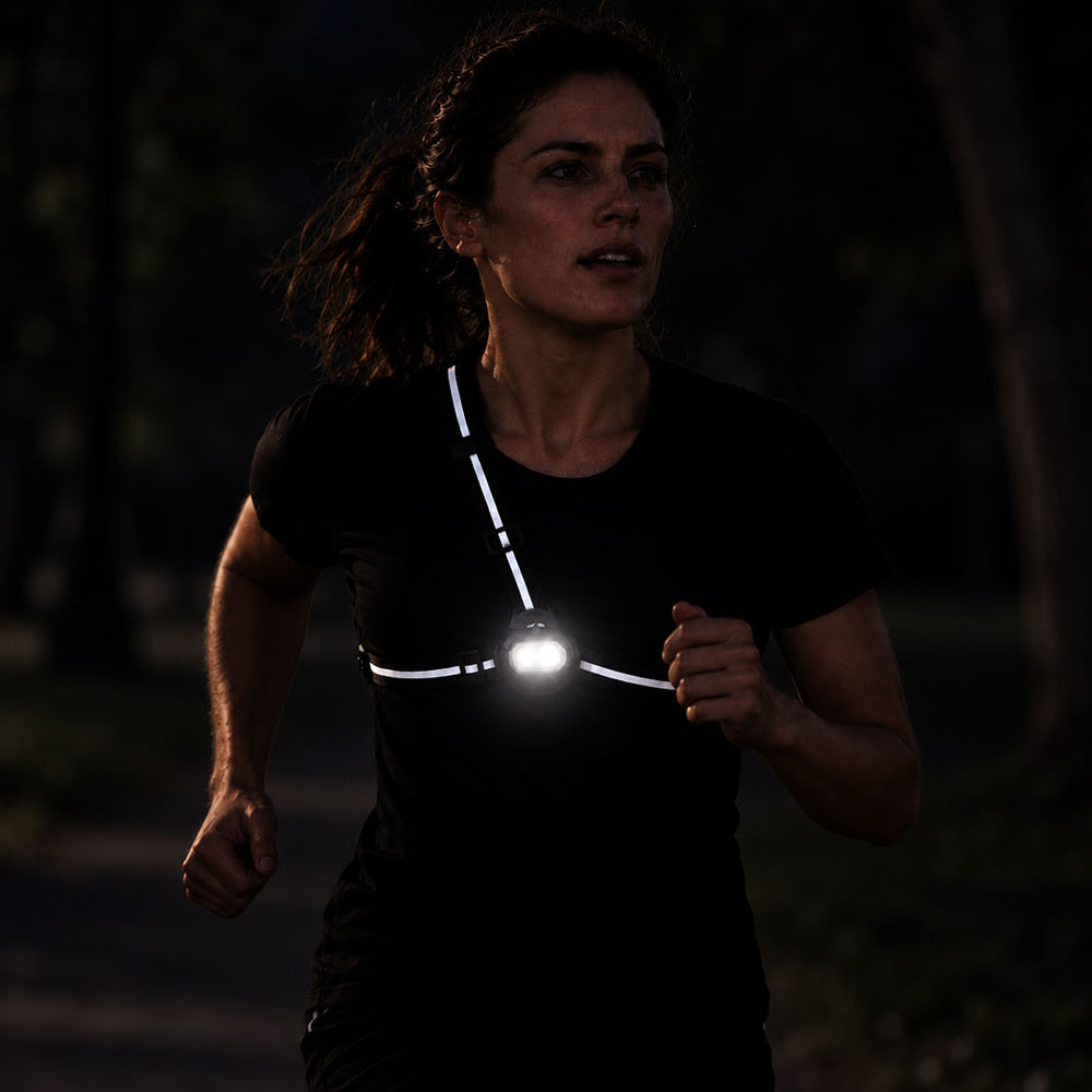 lifestyle A woman running outdoors at night wearing a reflective  proviz chest strap light with a bright front bulb, shown against a dark blurred background.