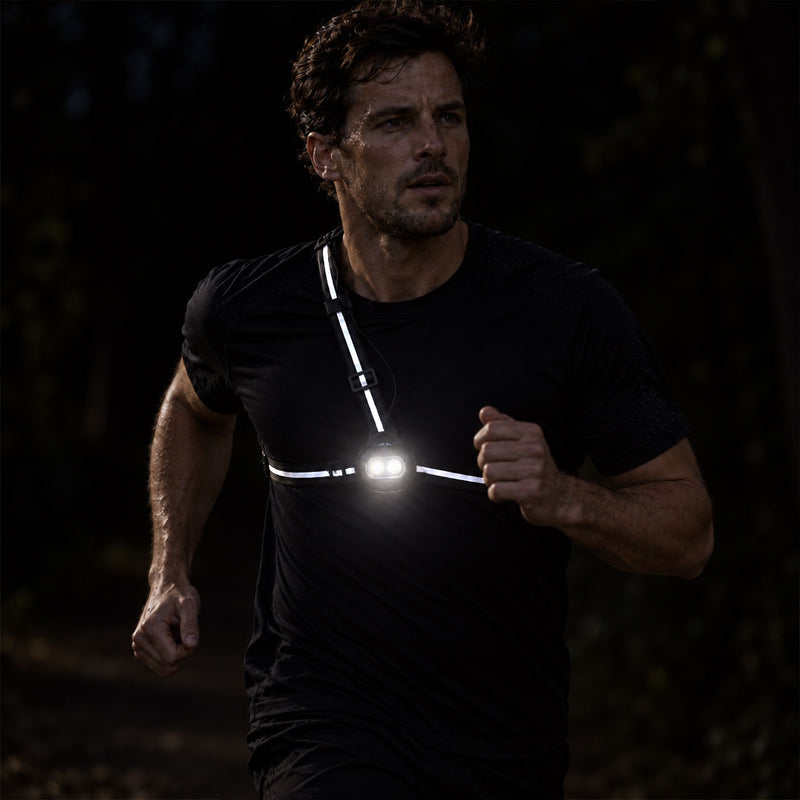lifestyle A man running outdoors at night wearing a black athletic shirt with reflective proviz chest straps and a bright front light, shown in a dark forest setting.