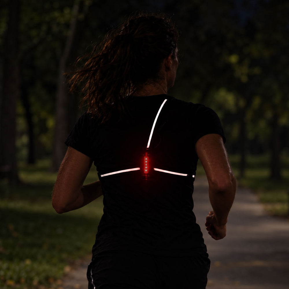 lifestyle A woman running on a dark park path viewed from behind, wearing a black shirt with glowing white reflective back stripes and a small proviz red light in the center.