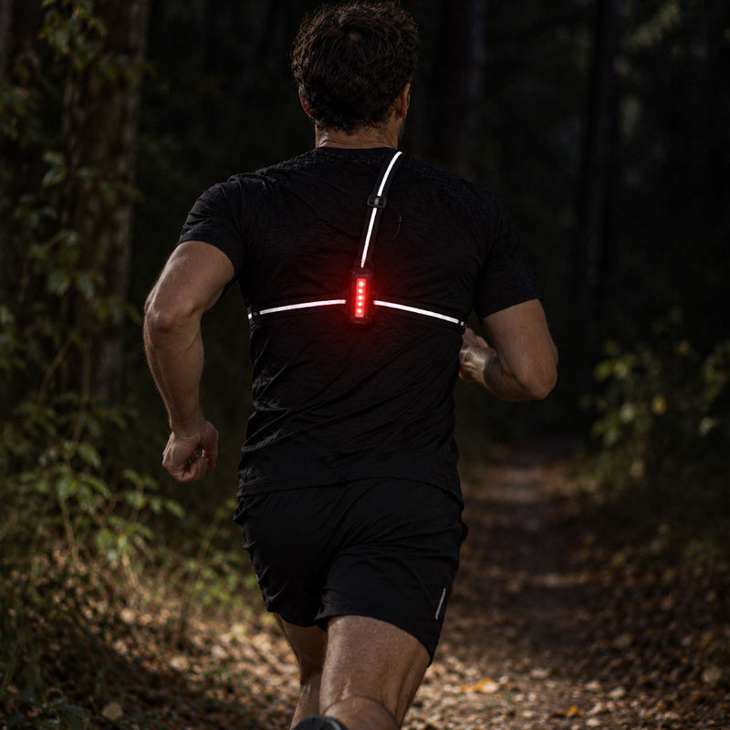 lifestyle A man running on a forest path viewed from behind, wearing a black athletic shirt and shorts with a red rear light and proviz white reflective-looking strap details on the back.