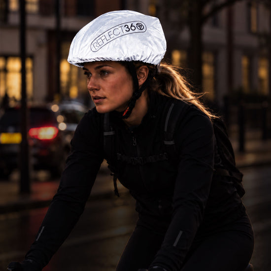 listing-lifestyle A cyclist wearing a reflective silver proviz helmet cover with the brand name 'REFLECT360' printed on it, riding on a city street at night.