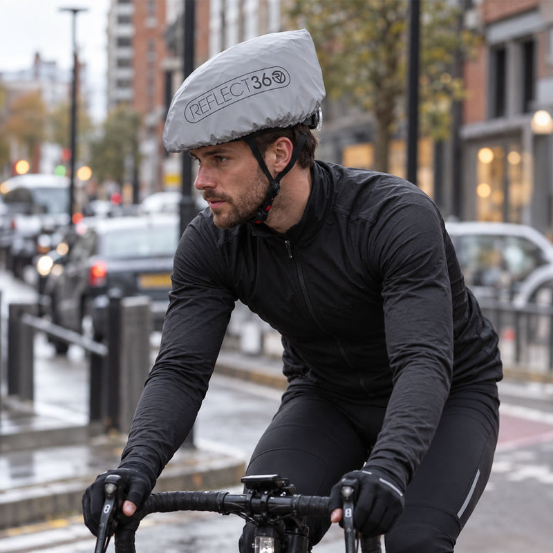 lifestyle A man riding a bicycle on a city street wearing a reflective gray proviz helmet cover with the brand name 'REFLECT360' printed on the front, along with a black cycling jersey and black gloves.