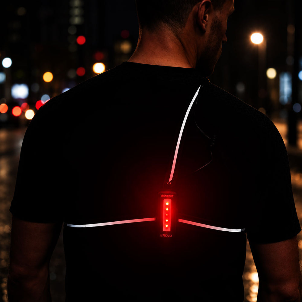 lifestyle A man viewed from behind at night wearing a black t-shirt with reflective white lines and a glowing red rear light bar. The background shows a blurred city street at night with warm lights. Man is wearign listing-lifestyle Running Chest Light at 500 Lumens outside light on