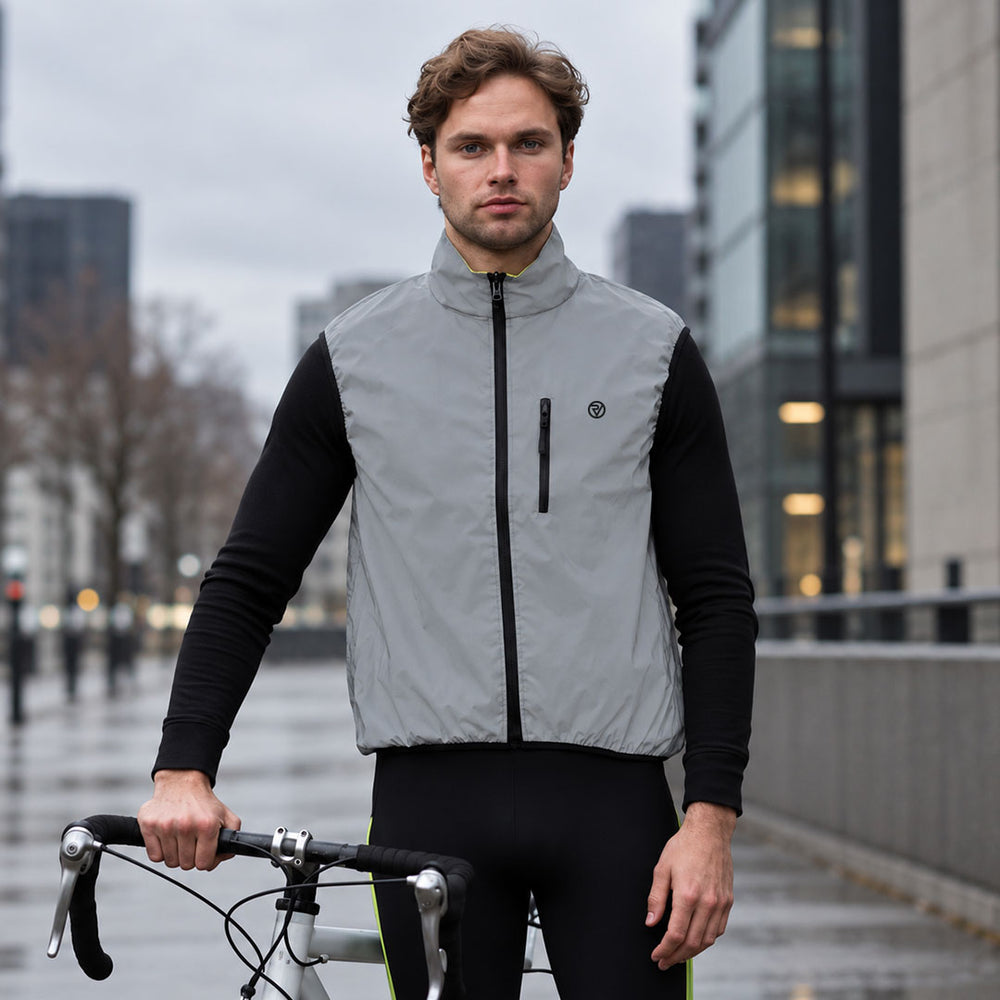lifestyle A man wearing a Proviz light gray cycling vest over a black long-sleeve shirt and black cycling tights, standing on a city street with a bicycle handlebar visible in the foreground.