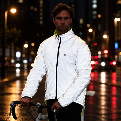 lifestyle A man wearing a Proviz white reflective cycling jacket with a black zipper and a small circular logo on the chest stands on a wet city street at night, holding a bicycle.
