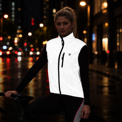 listing-lifestyle A woman wearing a Proviz white reflective cycling vest over a black long-sleeve top and black leggings, posing on a bicycle in a wet city street at night with blurred streetlights in the background.