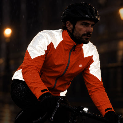 lifestyle A man riding a bicycle in the rain at night wearing a bright orange and white Proviz waterproof cycling jacket, black helmet, and black pants.