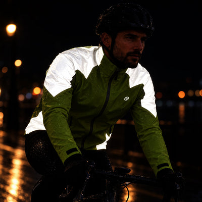 listing-lifestyle A man riding a bicycle at night in the rain, wearing a green and white waterproof  proviz cycling jacket, black helmet, and black gloves. The background is dark with blurred streetlights reflecting on wet pavement.