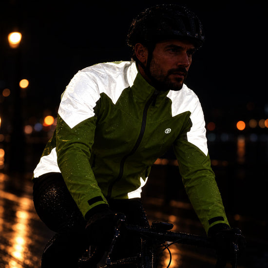 listing-lifestyle A man riding a bicycle at night in the rain, wearing a green and white waterproof  proviz cycling jacket, black helmet, and black gloves. The background is dark with blurred streetlights reflecting on wet pavement.
