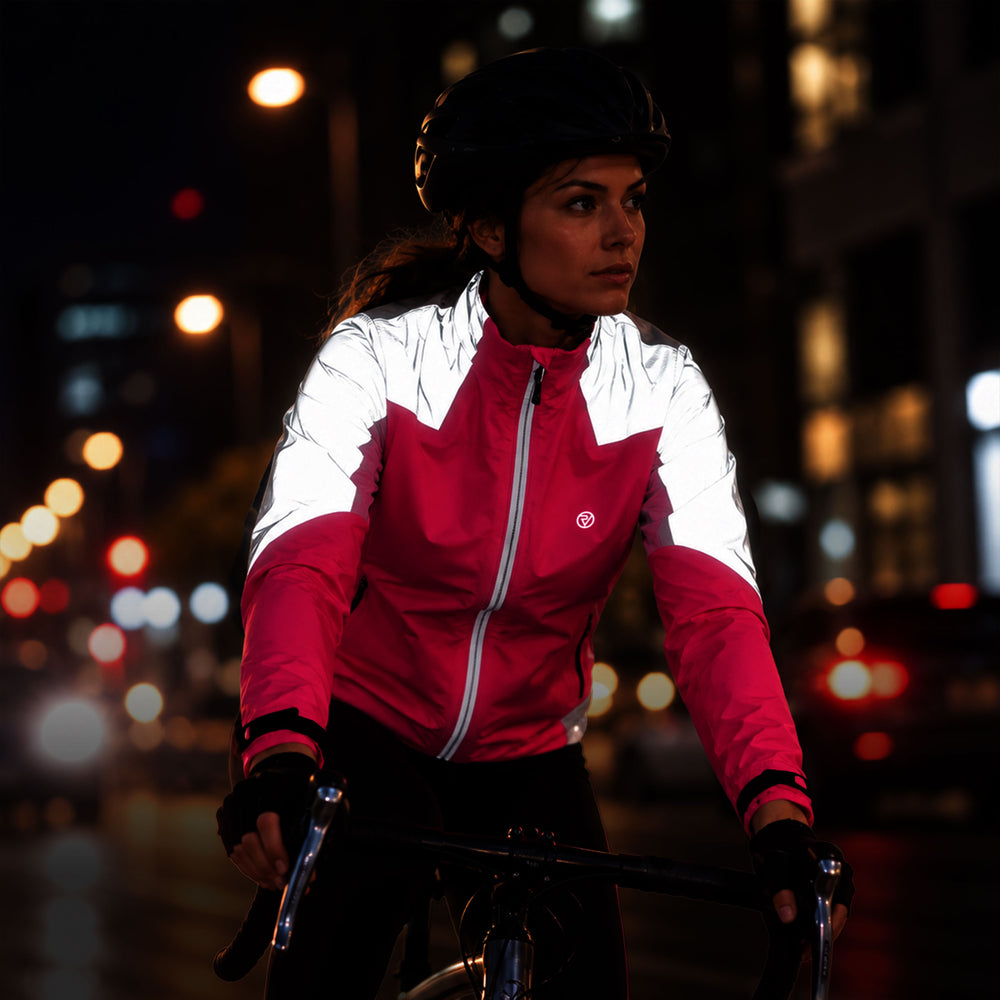 lifestyle A woman wearing a Proviz reflective cycling jacket, black helmet, and gloves rides a bicycle at night on a city street with blurred streetlights in the background.