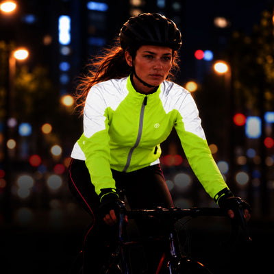lifestyle A woman wearing a Proviz reflective cycling jacket in neon yellow-green and white, along with a black helmet and gloves, riding a bicycle at night with blurred city lights in the background.