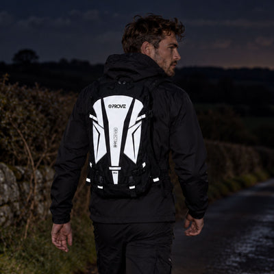 lifestyle A man wearing a black jacket and black pants walks outdoors on a dimly lit path, viewed from behind to show a black and reflective Proviz hydration pack on his back.