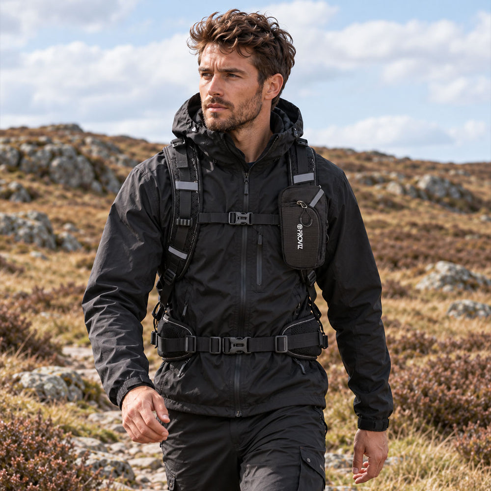 lifestyle A man hiking outdoors wearing a black hooded jacket with a built-in hydration pack and a black pouch on the chest that reads 'Proviz'.