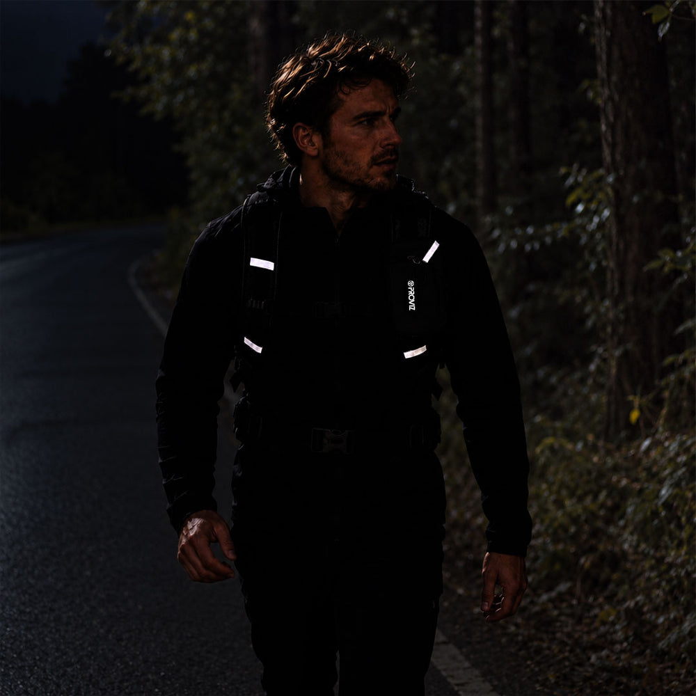 lifestyle A man walking on a dark road at night wearing a black reflective proviz backpack with white reflective strips and a black hoodie, with trees on the side of the road.