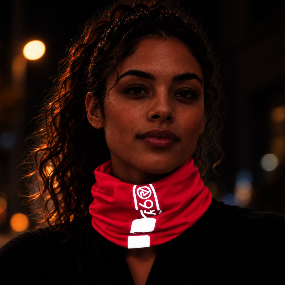 lifestyle A close-up portrait of a woman wearing a Proviz bright pink neck gaiter with reflective white striping and a white logo, set against a dark, softly blurred nighttime background with warm lights.
