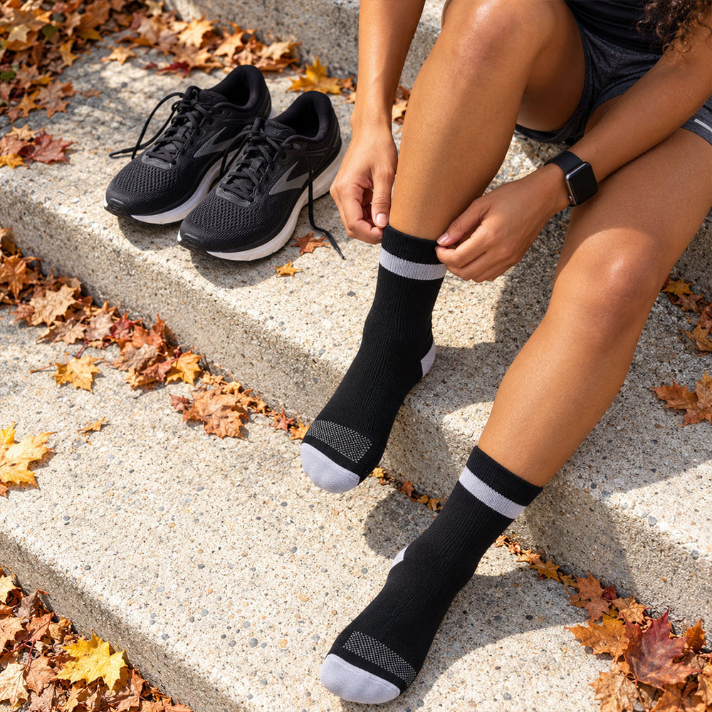 lifestyle A person sitting on concrete steps outdoors adjusts a Proviz black crew sock with a light gray band, with matching black athletic shoes nearby and fallen autumn leaves around them.