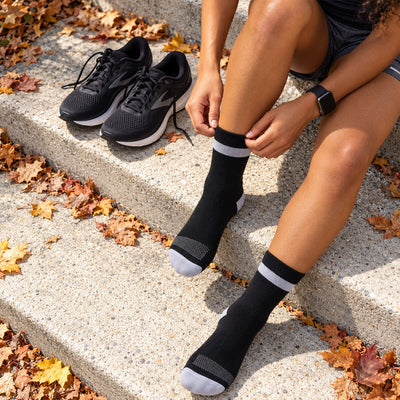 lifestyle A person sitting on concrete steps outdoors adjusts a Proviz black crew sock with a light gray band, with matching black athletic shoes nearby and fallen autumn leaves around them.