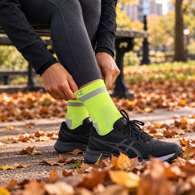lifestyle A person sitting on a bench in a park, adjusting a pair of bright neon yellow Proviz athletic socks with reflective gray stripes. The person is wearing black running shoes and dark gray leggings, with fallen autumn leaves covering the ground.