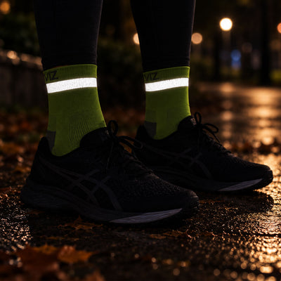 lifestyle A close-up of a person's legs wearing black running shoes and bright lime green reflective Proviz socks on a wet, reflective pavement at night.