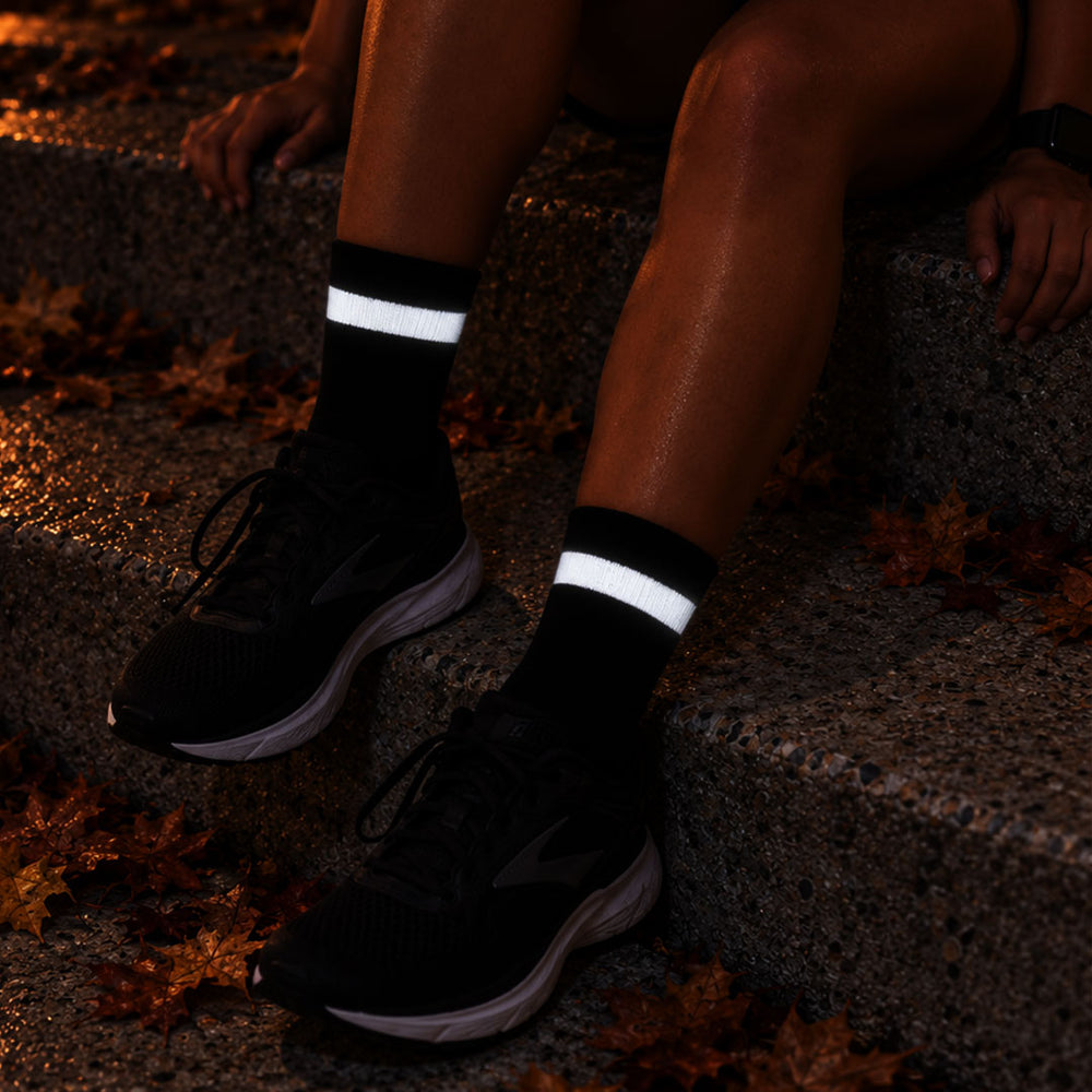 lifestyle Proviz Black athletic socks with reflective white horizontal stripes worn with black running shoes, shown on a person sitting on outdoor stone steps with fallen leaves.