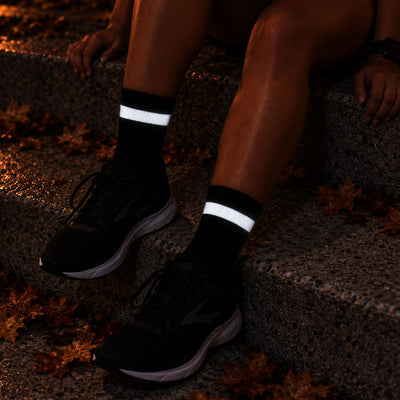 lifestyle Proviz Black athletic socks with reflective white horizontal stripes worn with black running shoes, shown on a person sitting on outdoor stone steps with fallen leaves.
