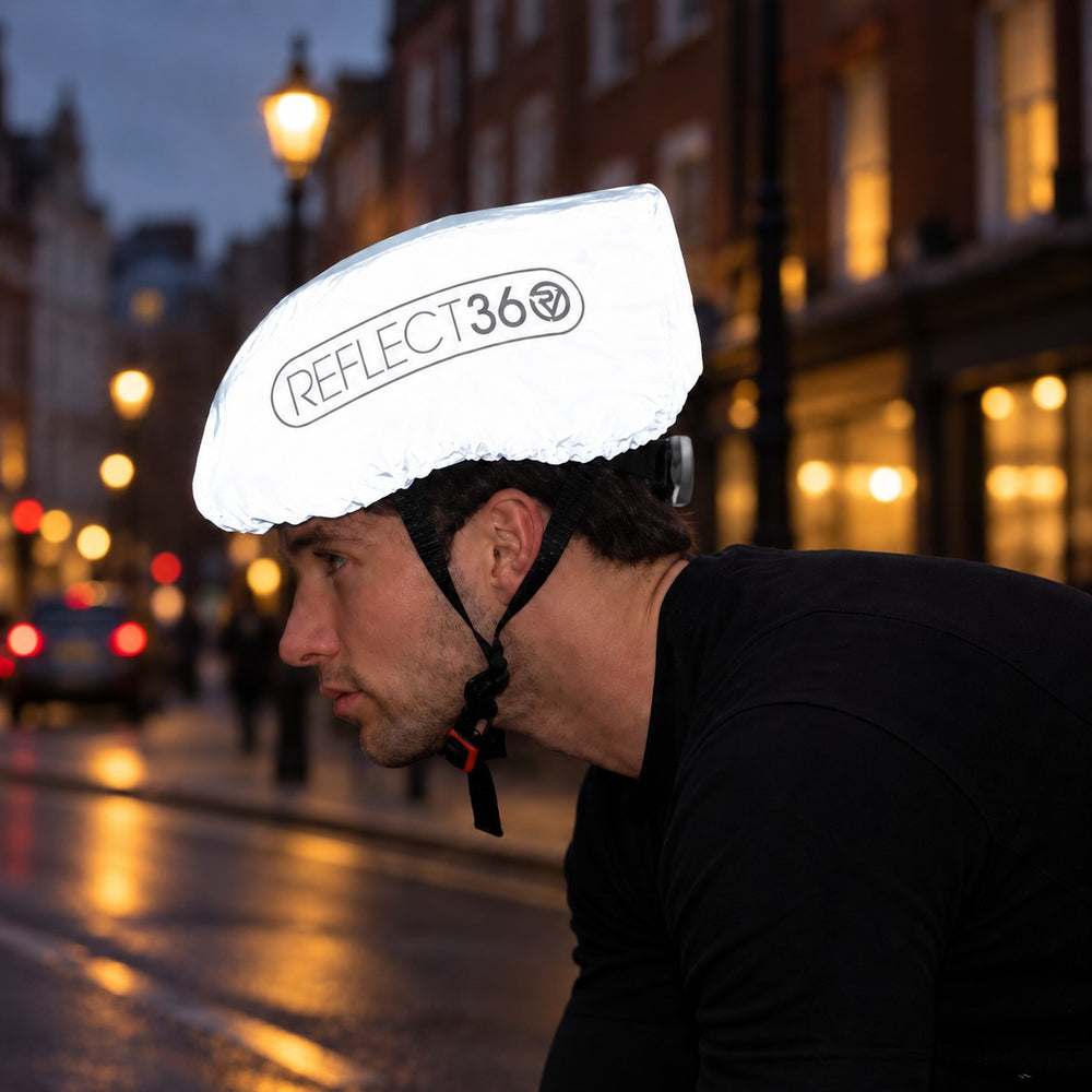 lifestyle A man wearing a Proviz  reflective white bicycle helmet cover with the brand name 'REFLECT36' printed on the front, riding on a city street at dusk.