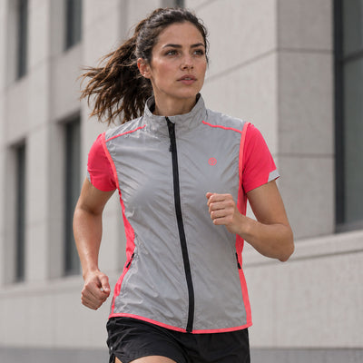 lifestyle A woman running outdoors wearing a Proviz reflective and bright pink athletic zip-up vest with black shorts, set against a blurred modern building background.