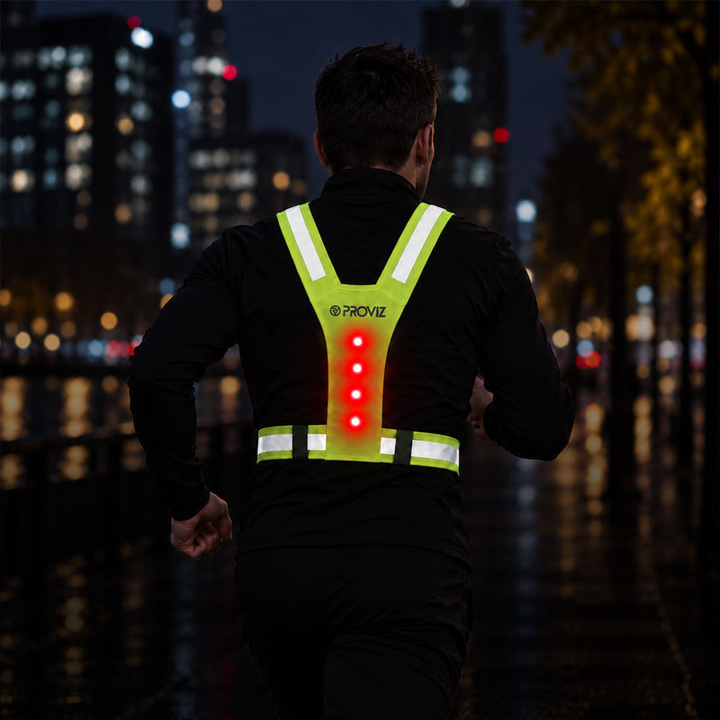 lifestyle A man running at night wearing a reflective Proviz running vest with illuminated red LED lights on the back, set against a blurred city street background.
