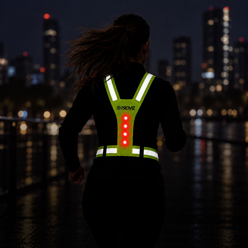 lifestyle A woman running at night from behind on a city waterfront, wearing a black long-sleeve top with a bright green reflective vest featuring white reflective strips and glowing red LED lights on the back. The brand name 'PROVZ' is visible on the back of the vest.