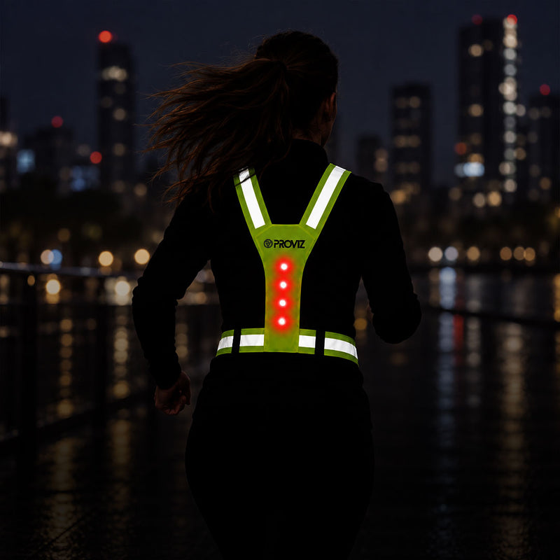 lifestyle A woman running at night from behind on a city waterfront, wearing a black long-sleeve top with a bright green reflective vest featuring white reflective strips and glowing red LED lights on the back. The brand name 'PROVZ' is visible on the back of the vest.