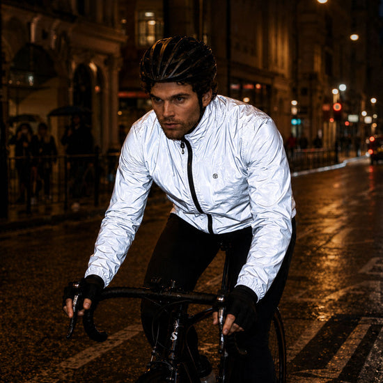 listing-lifestyle A cyclist riding on a wet city street at night, wearing a Proviz shiny light gray reflective cycling jacket, black helmet, and black gloves.