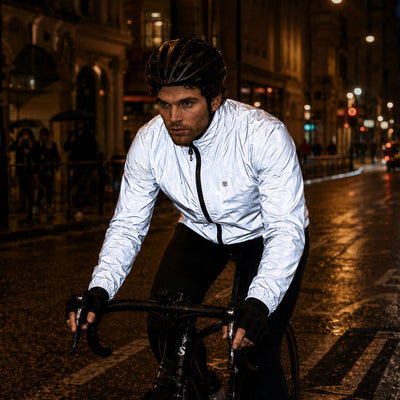 listing-lifestyle A cyclist riding on a wet city street at night, wearing a Proviz shiny light gray reflective cycling jacket, black helmet, and black gloves.
