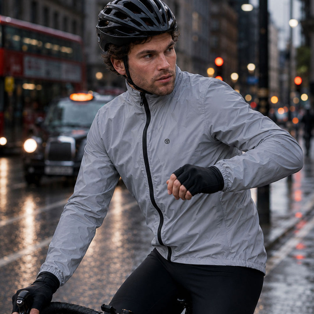 lifestyle A man wearing a Proviz light gray waterproof cycling jacket, black helmet, and black gloves rides a bicycle on a wet city street at night.