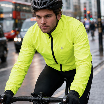 lifestyle A man wearing a Proviz bright neon yellow-green cycling jacket, black helmet, and black cycling tights rides a city bike on a wet street with blurred cars and a red double-decker bus in the background.