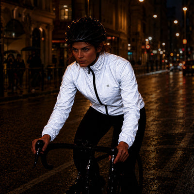 lifestyle A woman riding a bicycle at night on a wet city street, wearing a shiny white reflective proviz cycling jacket, black helmet, and black pants.