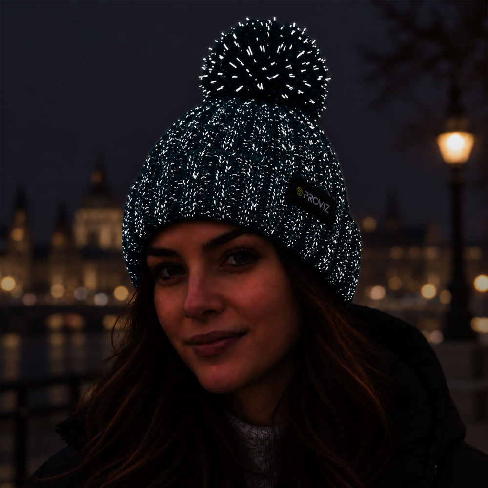 lifestyle A woman wearing a black knit beanie with reflective silver flecks and a black pom-pom, photographed outdoors at night with blurred city lights in the background. A small black brand tag on the side reads PROVIZ.