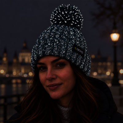 lifestyle A woman wearing a black knit beanie with reflective silver flecks and a black pom-pom, photographed outdoors at night with blurred city lights in the background. A small black brand tag on the side reads PROVIZ.