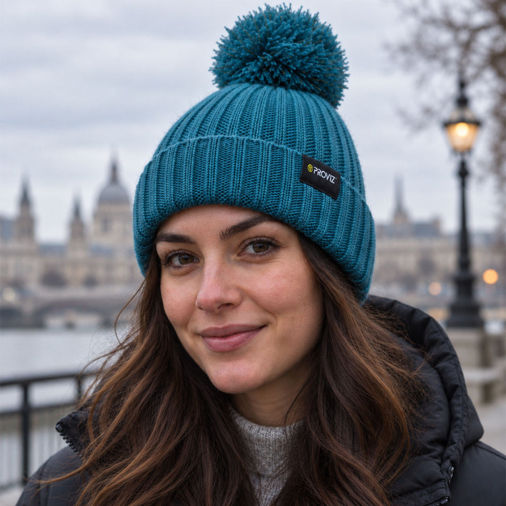 lifestyle A woman wearing a teal blue knit beanie with a matching pom-pom and a small black brand patch that reads 'Proviz'. The background shows a softly blurred outdoor city scene with a river and historic building.