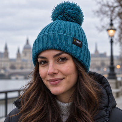 lifestyle A woman wearing a teal blue knit beanie with a matching pom-pom and a small black brand patch that reads 'Proviz'. The background shows a softly blurred outdoor city scene with a river and historic building.