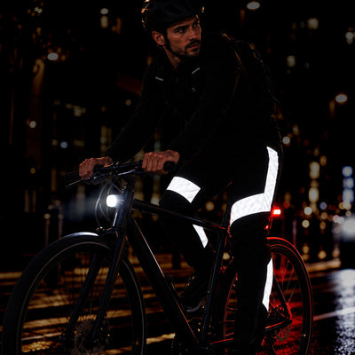 lifestyle A man riding a bicycle at night wearing reflective safety gear, including high-visibility white reflective bands on his pants and a black helmet, with a bicycle headlight illuminated on a wet city street.