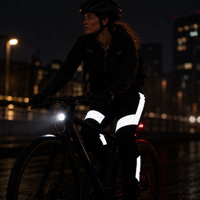 listing-lifestyle A woman riding a bicycle at night wearing reflective proviz cycling trousers and a black jacket, with her bike's headlight on and a blurred city skyline in the background.