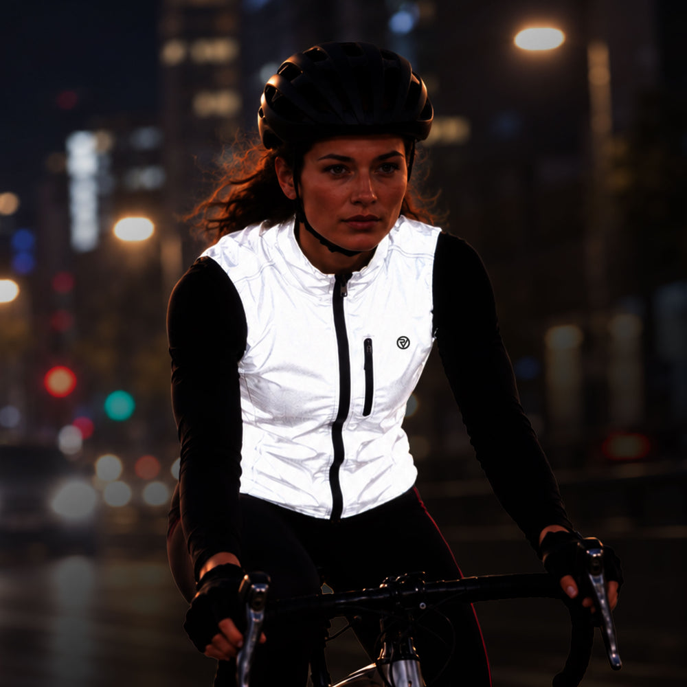 lifestyle A woman wearing a Proviz white reflective cycling vest over a black long-sleeve top and black cycling tights rides a bicycle at night on a city street with blurred streetlights in the background.