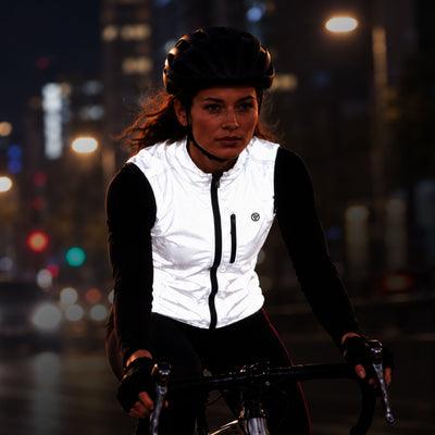 lifestyle A woman wearing a Proviz white reflective cycling vest over a black long-sleeve top and black cycling tights rides a bicycle at night on a city street with blurred streetlights in the background.