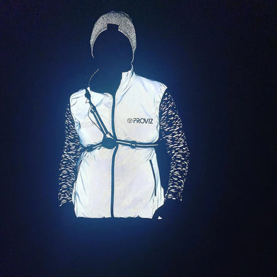 lifestyle A person wearing a reflective proviz white vest with black zippers and a black chest strap, paired with patterned sleeves and a reflective beanie, shown against a dark background.